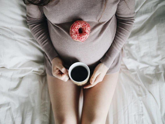 A pregnant woman sits on a bed with coffee and a doughnut